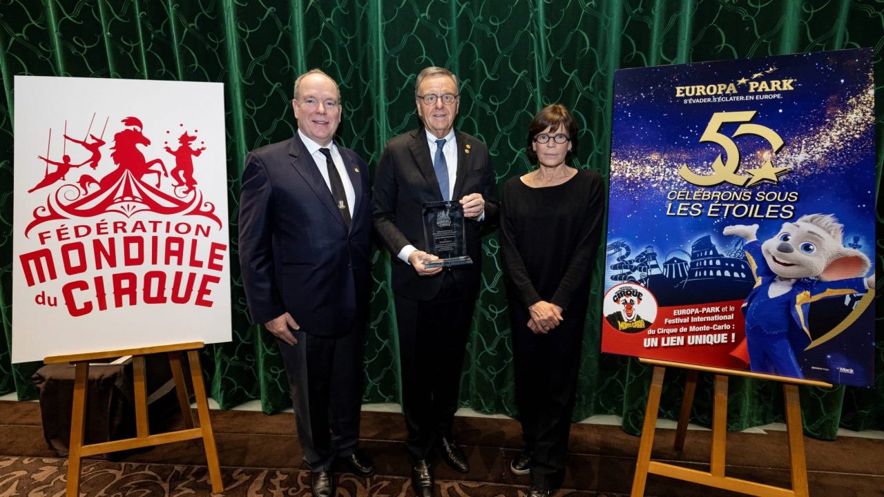 Drei Personen stehen vor einem grünen Vorhang. Links S.D. Fürst Albert II. von Monaco in dunklem Anzug mit Krawatte, in der Mitte Roland Mack, der eine Auszeichnung in den Händen hält, und rechts I.D. Prinzessin Stéphanie von Monaco in einem schwarzen Outfit. Links im Bild ein Aufsteller mit dem Logo der „Fédération Mondiale du Cirque“, rechts ein Plakat mit dem Europa-Park-Maskottchen und dem Text „50 – Célébrons sous les étoiles“.