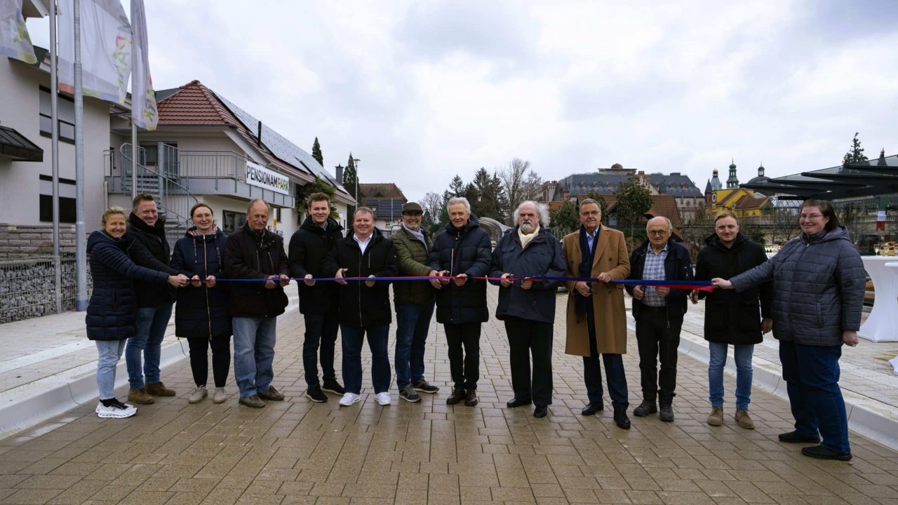 Eine Gruppe von Menschen steht in einer Reihe und hält ein Band, das für eine feierliche Eröffnung durchschnitten werden soll. Die Personen sind in warme Kleidung gehüllt, was auf kühles Wetter hindeutet. Im Hintergrund sind Gebäude mit traditioneller Architektur zu sehen, darunter ein Gebäude mit gelber Fassade und grünen Türmen sowie eine Pension mit Solarpanelen auf dem Dach. Rechts im Bild stehen Stehtische mit Getränken, was auf eine offizielle Veranstaltung hinweist.