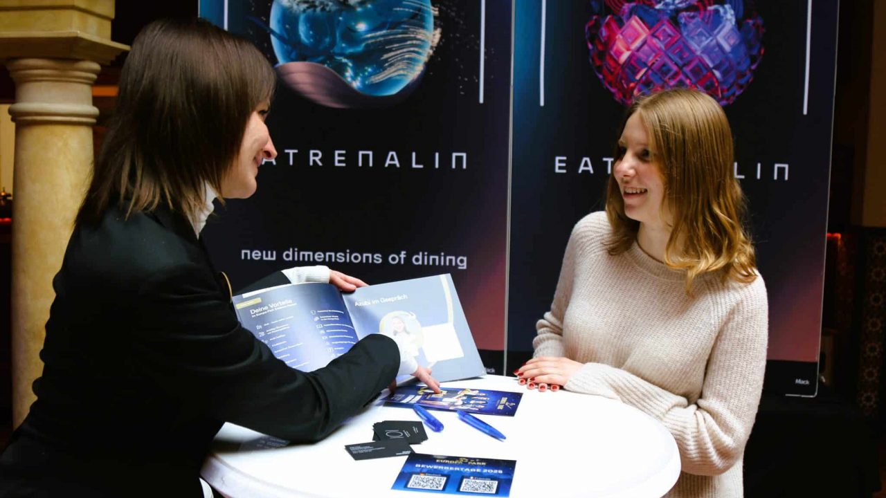 Zwei Personen bei einem Bewerbungsgespräch an einem runden Tisch. Eine Frau in schwarzem Blazer hält eine Broschüre aufgeschlagen und zeigt auf Informationen, während eine junge Frau in einem beigen Pullover ihr aufmerksam zuhört und lächelt. Im Hintergrund sind Plakate mit dem Schriftzug "EATRENALIN – new dimensions of dining" zu sehen. Der Tisch ist mit Flyern, Stiften und Visitenkarten bedeckt.