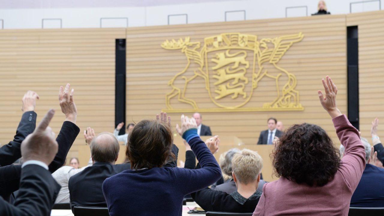Abgeordnete des Landtags von Baden-Württemberg heben in einer Plenarsitzung die Hände zur Abstimmung. Im Hintergrund ist das Landeswappen Baden-Württembergs zu sehen, während Redner an einem Podium stehen.