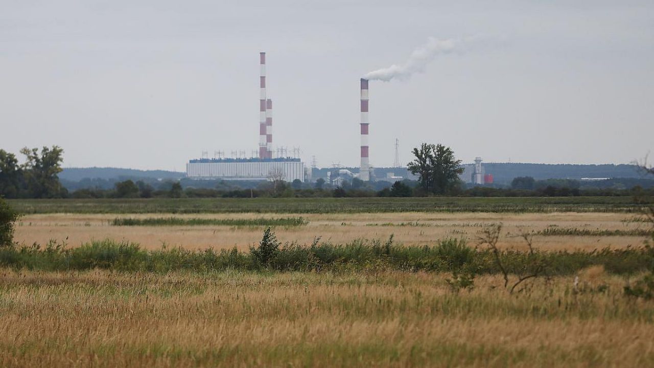 Industrieanlage mit hohen Schornsteinen und Rauch im Hintergrund, umgeben von Feldern.