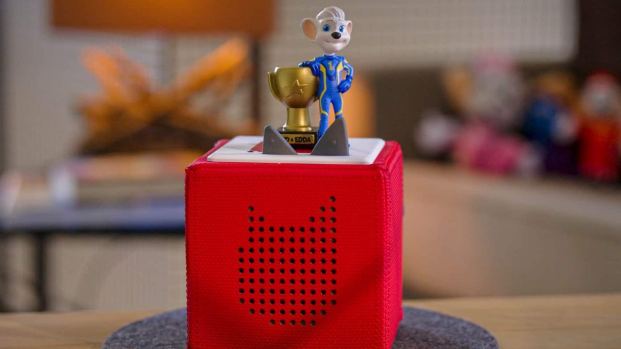 Ein rotes Toniebox-Hörspielgerät steht auf einem runden Filzuntersetzer auf einem Holztisch. Auf der Box befindet sich die Tonie-Figur Ed Euromaus, bekannt aus dem Europa-Park, in einem blauen Rennanzug, die eine goldene Trophäe hält. Im Hintergrund ist ein Wohnzimmer mit einer Lampe und verschwommenen Spielzeugfiguren zu sehen.